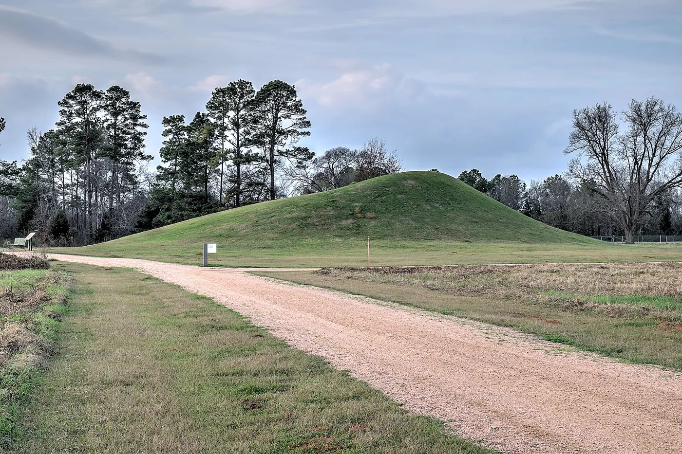 A grass-covered earthen mound beside a gravel path, with pine trees and a cloudy sky behind