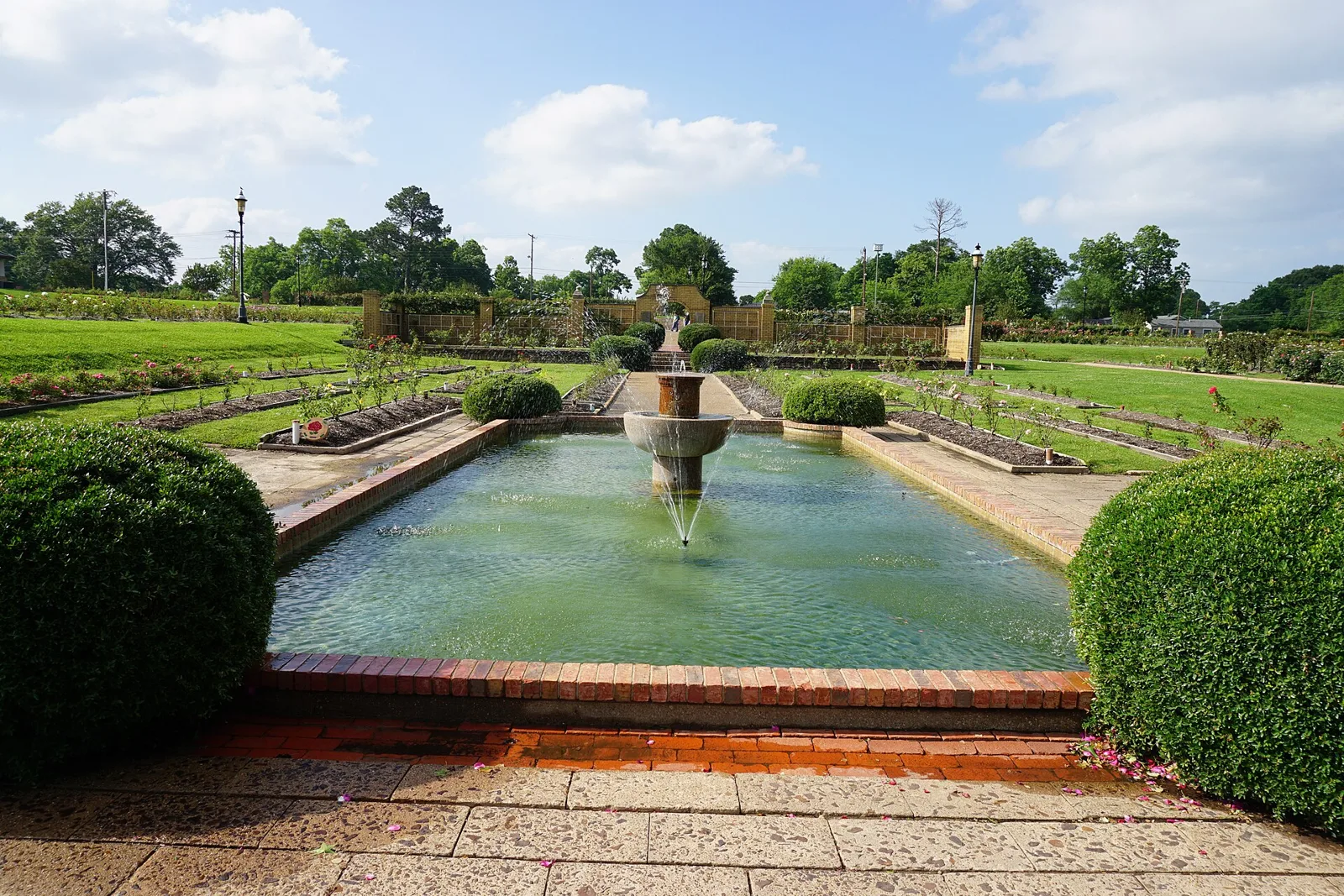 A long rectangular reflecting pool with a tiered fountain, framed by round boxwood shrubs, with rose beds stretching behind it